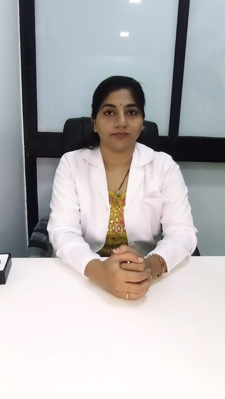 Dr. Rutuja Chapalgaokar a Female doctor in a white coat sitting at a clinic desk with hands folded, looking directly at the camera in a professional setting.